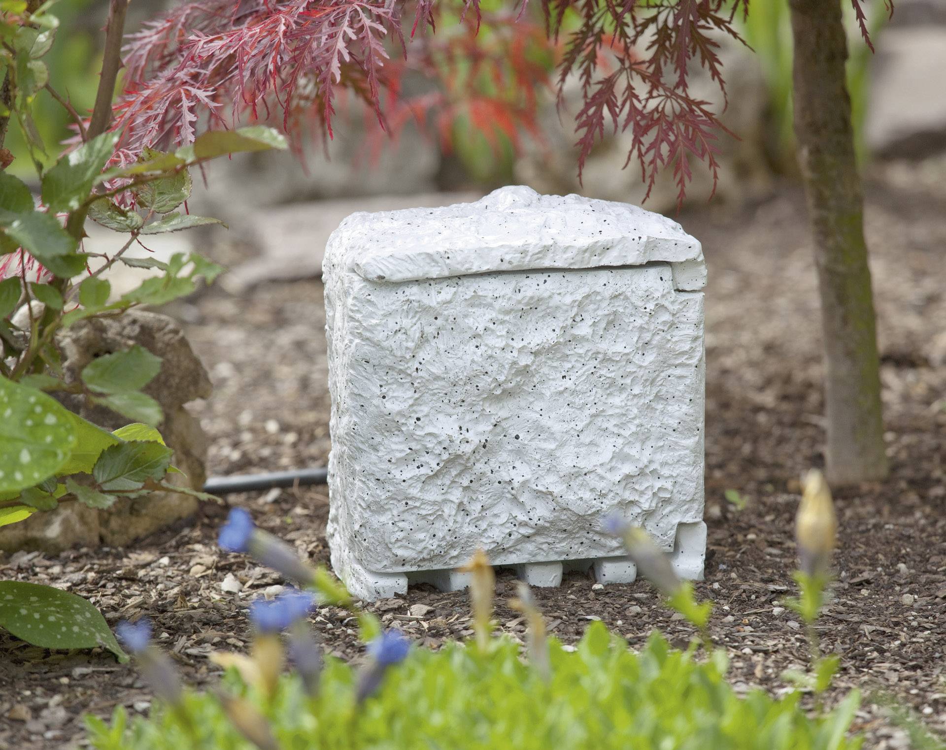 A white, cuboid stone speaker stands in a blooming garden, surrounded by plants and earth.
