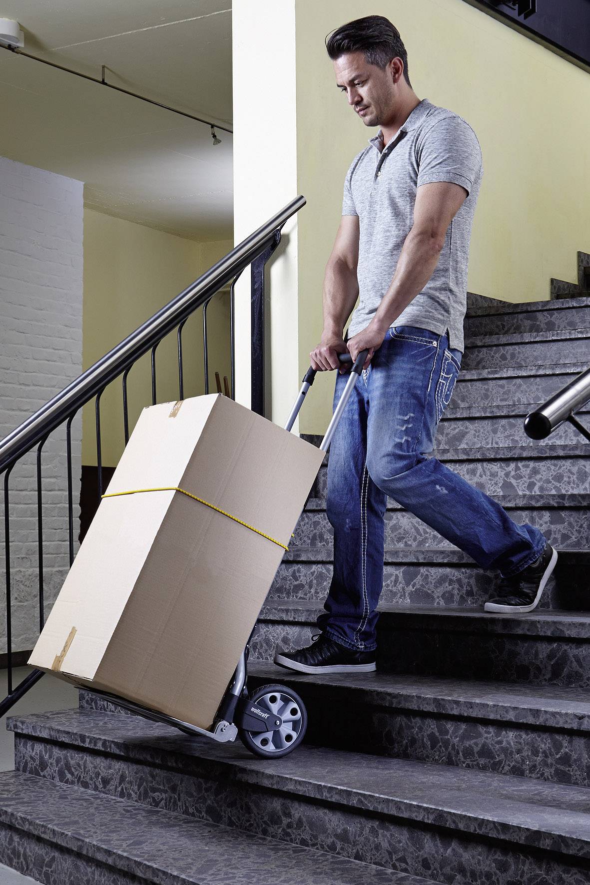 A man is carrying a large, brown cardboard box down some stairs using a hand trolley. He has a relaxed posture and is wearing casual clothing.