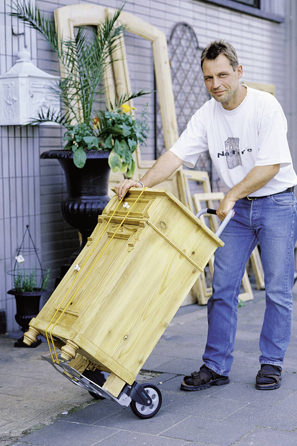 A man is pushing a yellow piece of furniture on a trolley. Plants and wooden frames can be seen in the background.