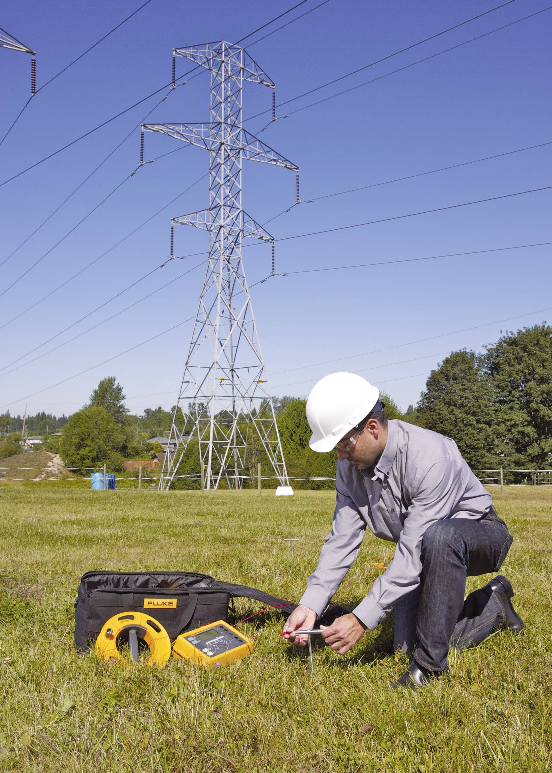 A worker wearing a hard hat is measuring electrical voltage with a measuring device in a field. A power transmission tower is visible in the background.