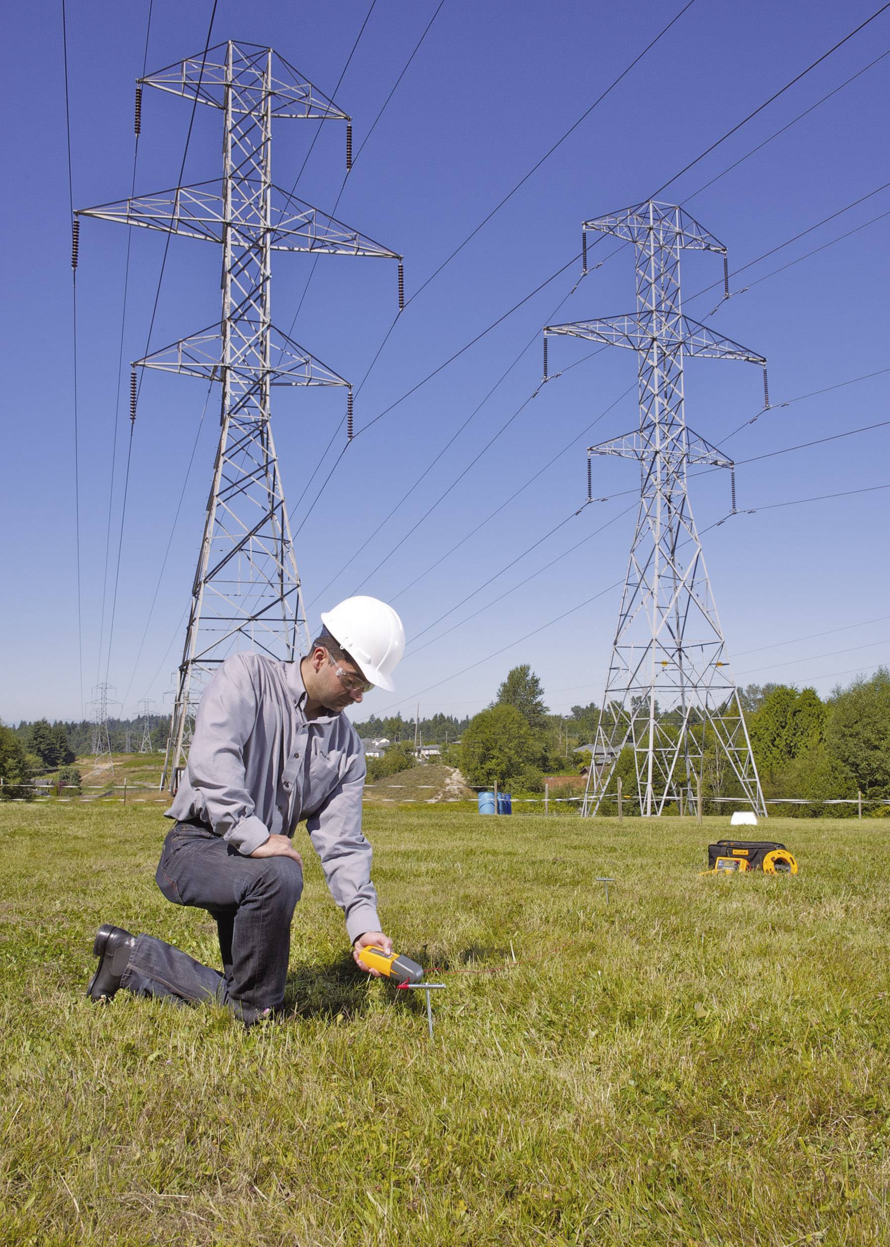 A technician wearing a hard hat is checking the earthing with a measuring device, with electricity pylons visible in a field in the background.