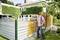 A man wearing a protective mask is painting a wooden fence with a spray gun in front of a house covered in ivy.