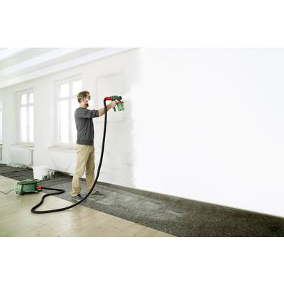 A man is spraying white paint with a spray gun onto a large, empty wall in a brightly lit room.