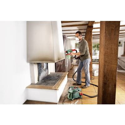 A man is wearing a respiratory mask and spraying paint onto a chimney with a paint sprayer in a room with wooden beam ceiling.