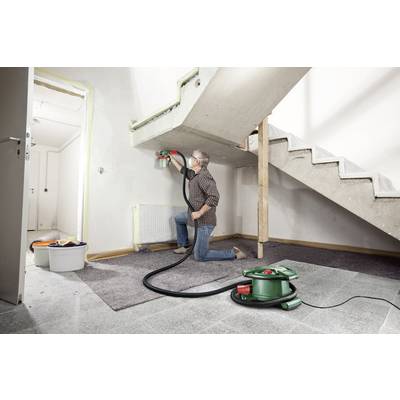 A man is kneeling on the floor and painting the wall under a staircase with a spray device. A bucket and a vacuum cleaner are visible.