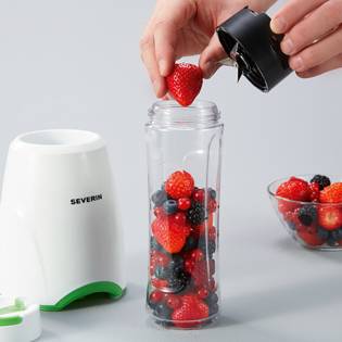 A person is placing a strawberry into a small blender that is already filled with berries. A bowl of berries is on the right, with the blender base on the left.