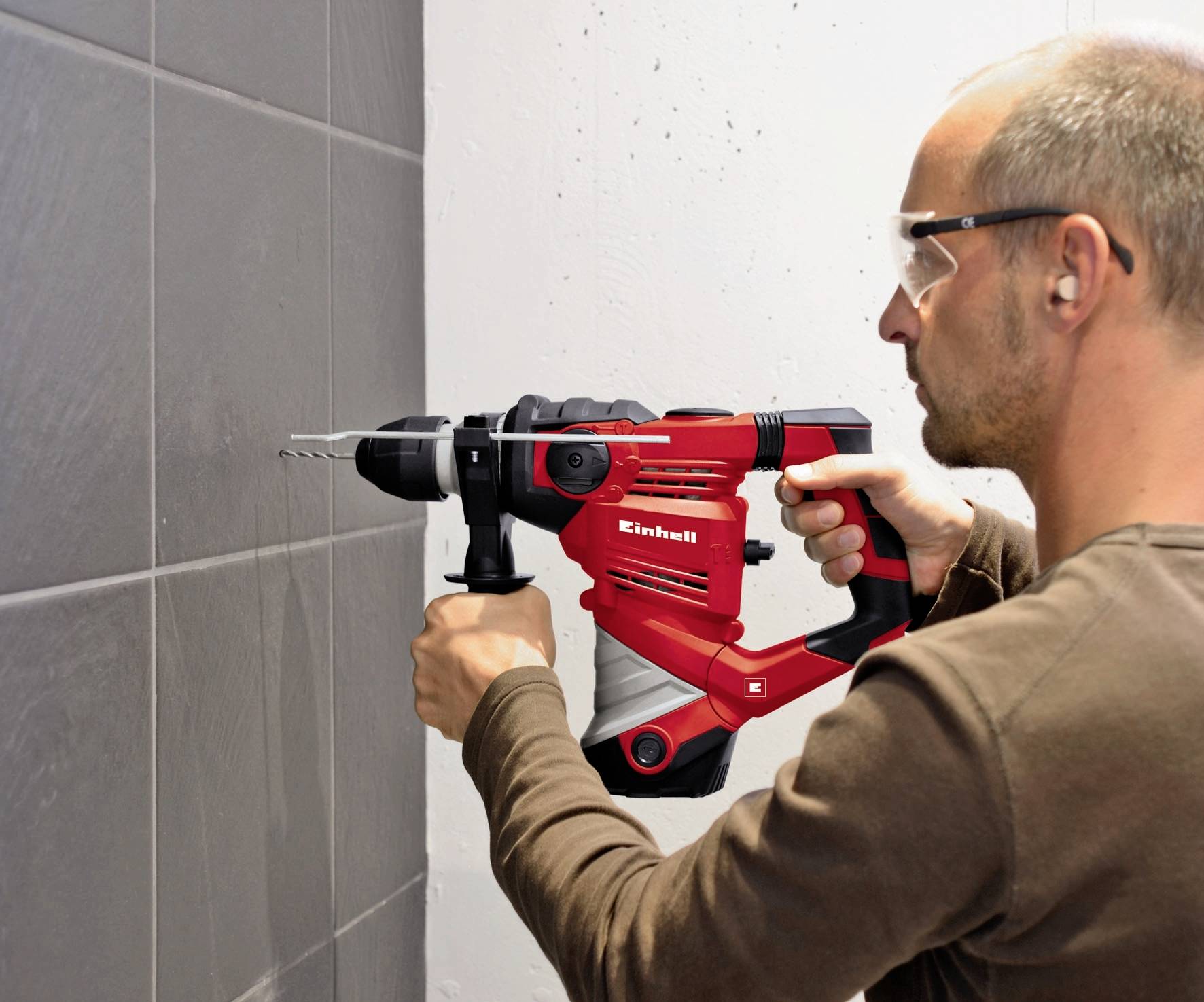 A man is wearing safety glasses and using a red drill hammer to bore a hole into a grey tile on the wall.