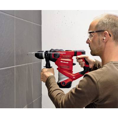 A man is wearing safety glasses and using a red drill hammer to bore a hole into a grey tile on the wall.
