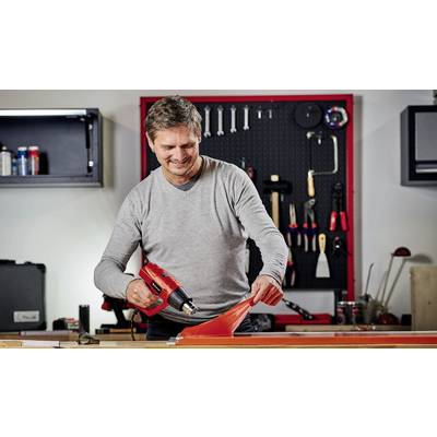 A man is working on skiing equipment with a hot air gun in a workshop. The background shows tools hanging on the wall.