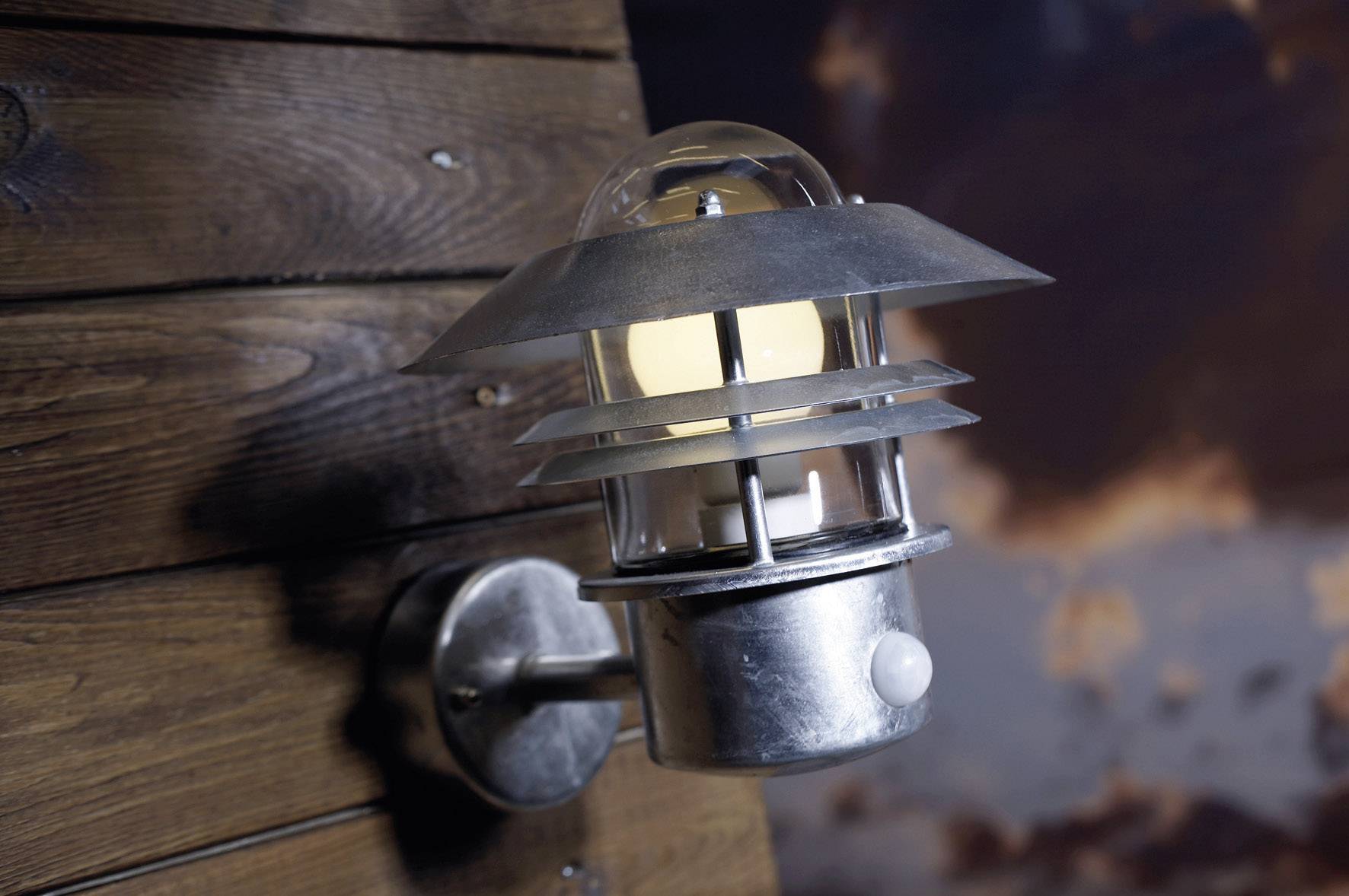 Metal outdoor lamp mounted on a wooden wall, illuminating at dusk. Background shows clouds with an evening atmosphere.