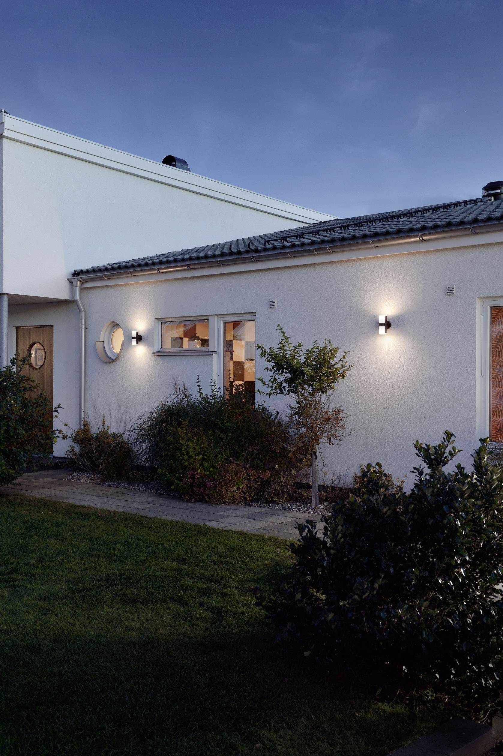 A modern detached house at dusk, illuminated by exterior lights. Shrubs and a well-maintained lawn surround the building.