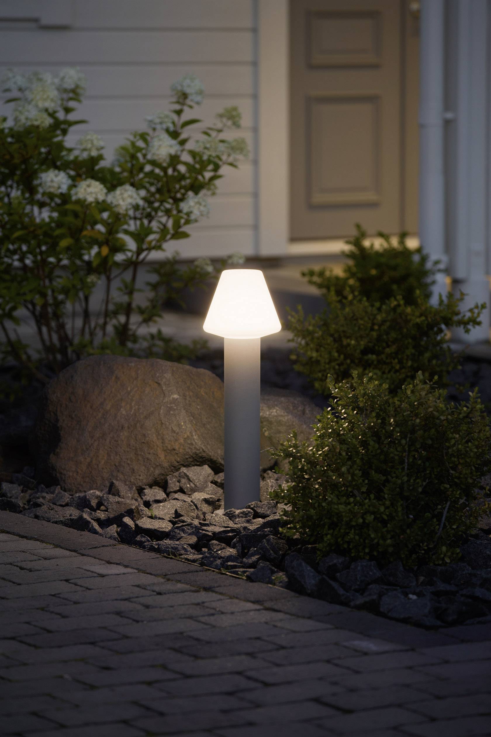'A luminous garden lamp beside a path, surrounded by plants and stones, in front of a house at dusk.'