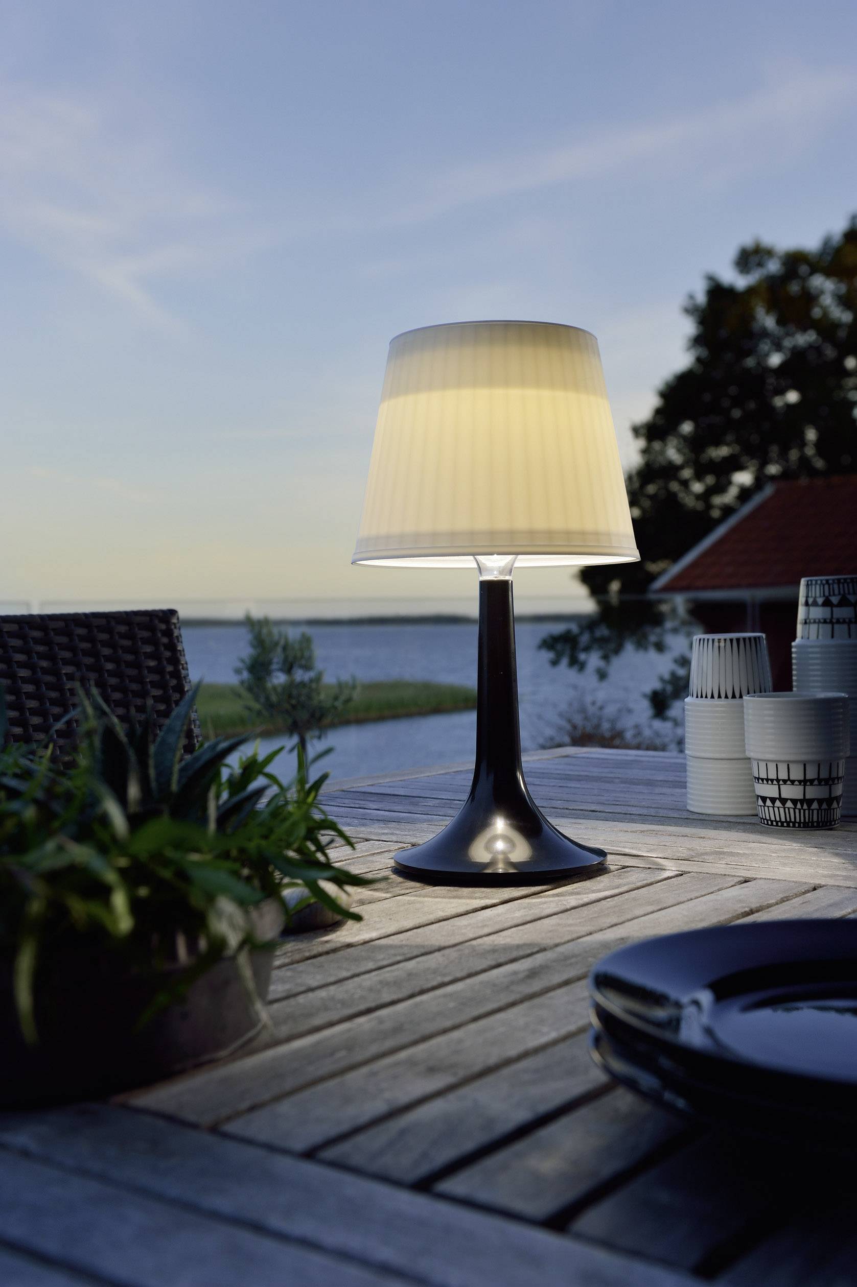 A table lamp illuminates a wooden table on a terrace in the evening. A lake and trees are visible in the background.