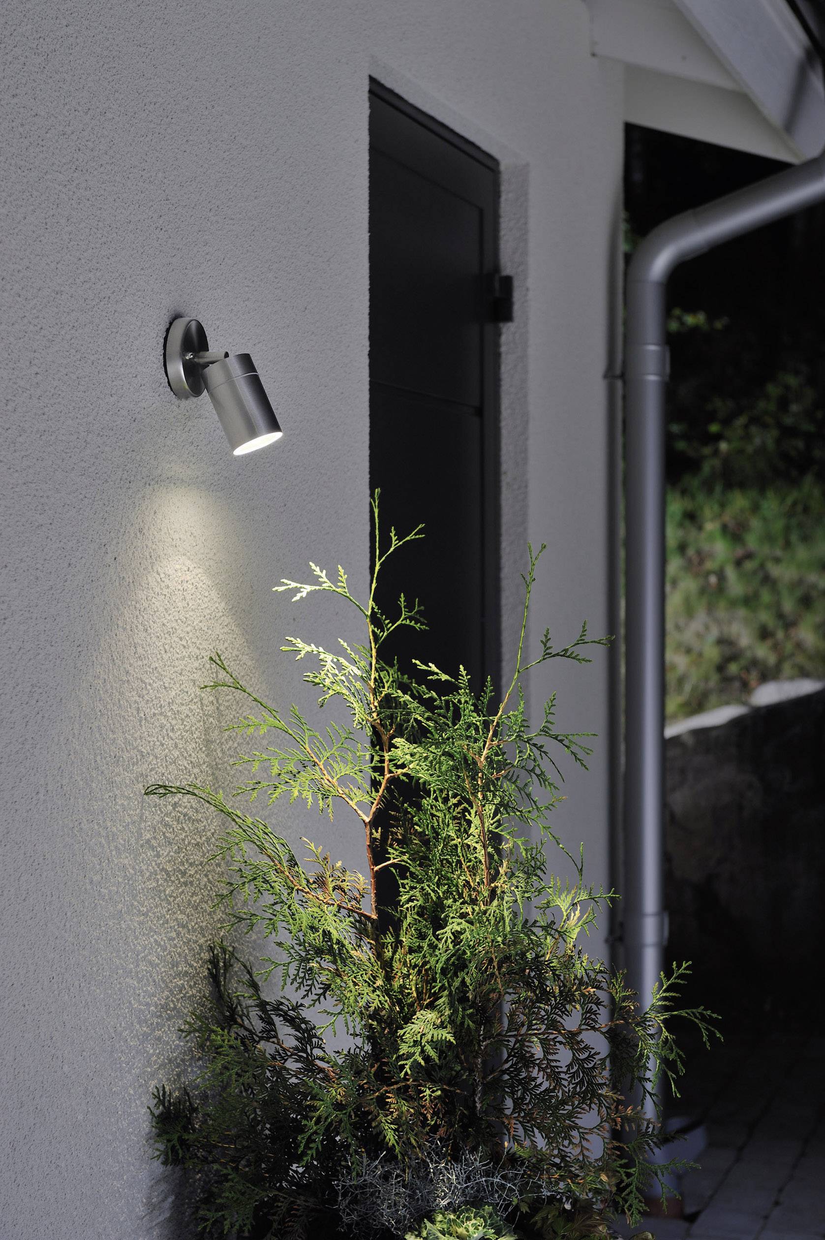 External lighting illuminates a house wall, beside a door and a green plant. Next to the wall is a downspout.