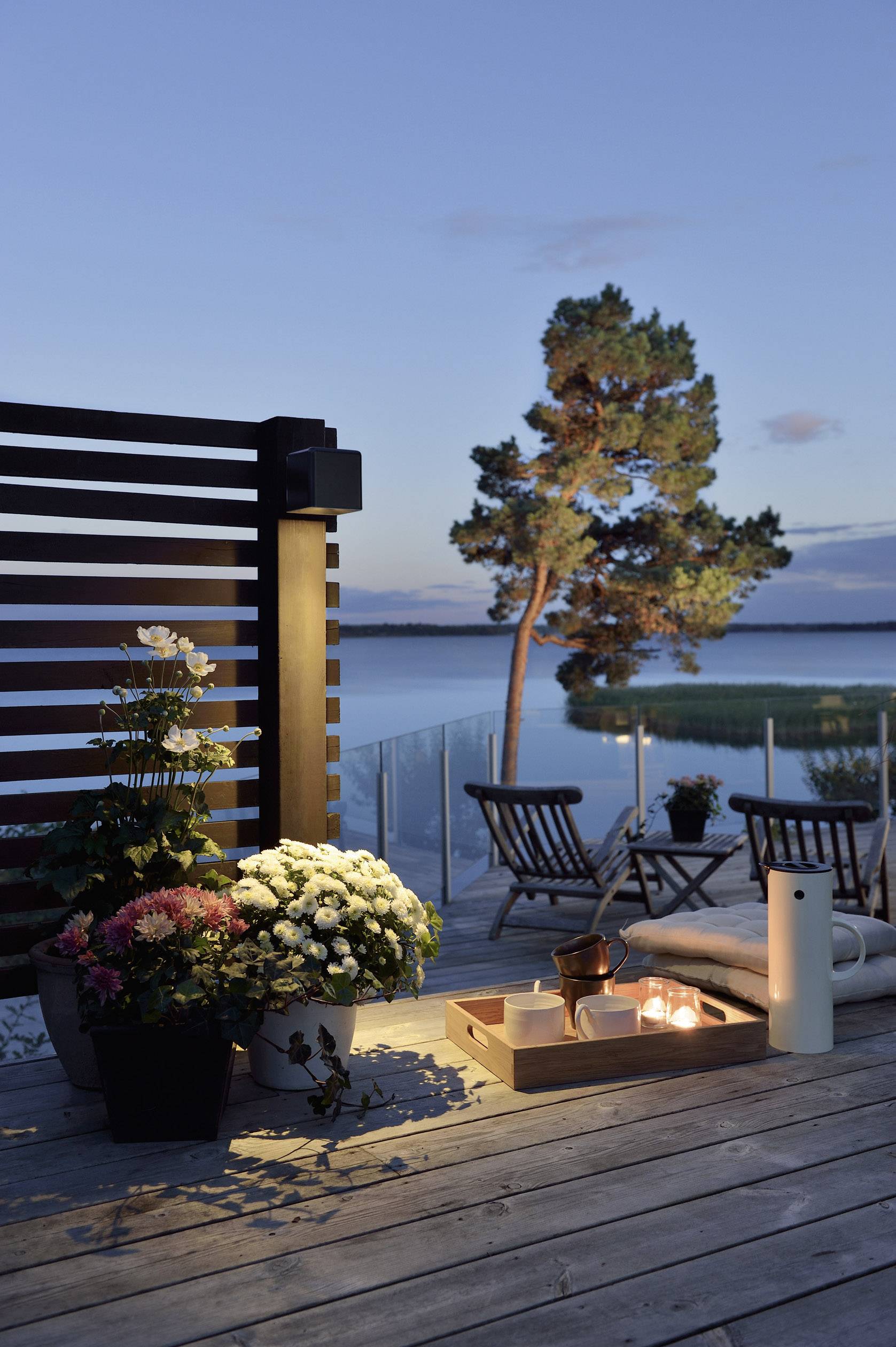 Terrace at sunset with candles on a wooden table, plants and two empty loungers overlooking a tranquil lake.