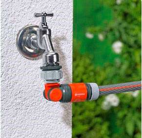 A tap on a white wall with a garden hose connected and extending to the right. Green vegetation is visible in the background.