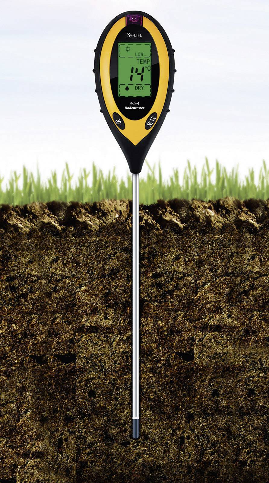 A digital soil measurement device is inserted into the ground and displays soil moisture, pH level, and temperature on its display. Grass blades in the background.