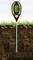 A digital soil measurement device is inserted into the ground and displays soil moisture, pH level, and temperature on its display. Grass blades in the background.