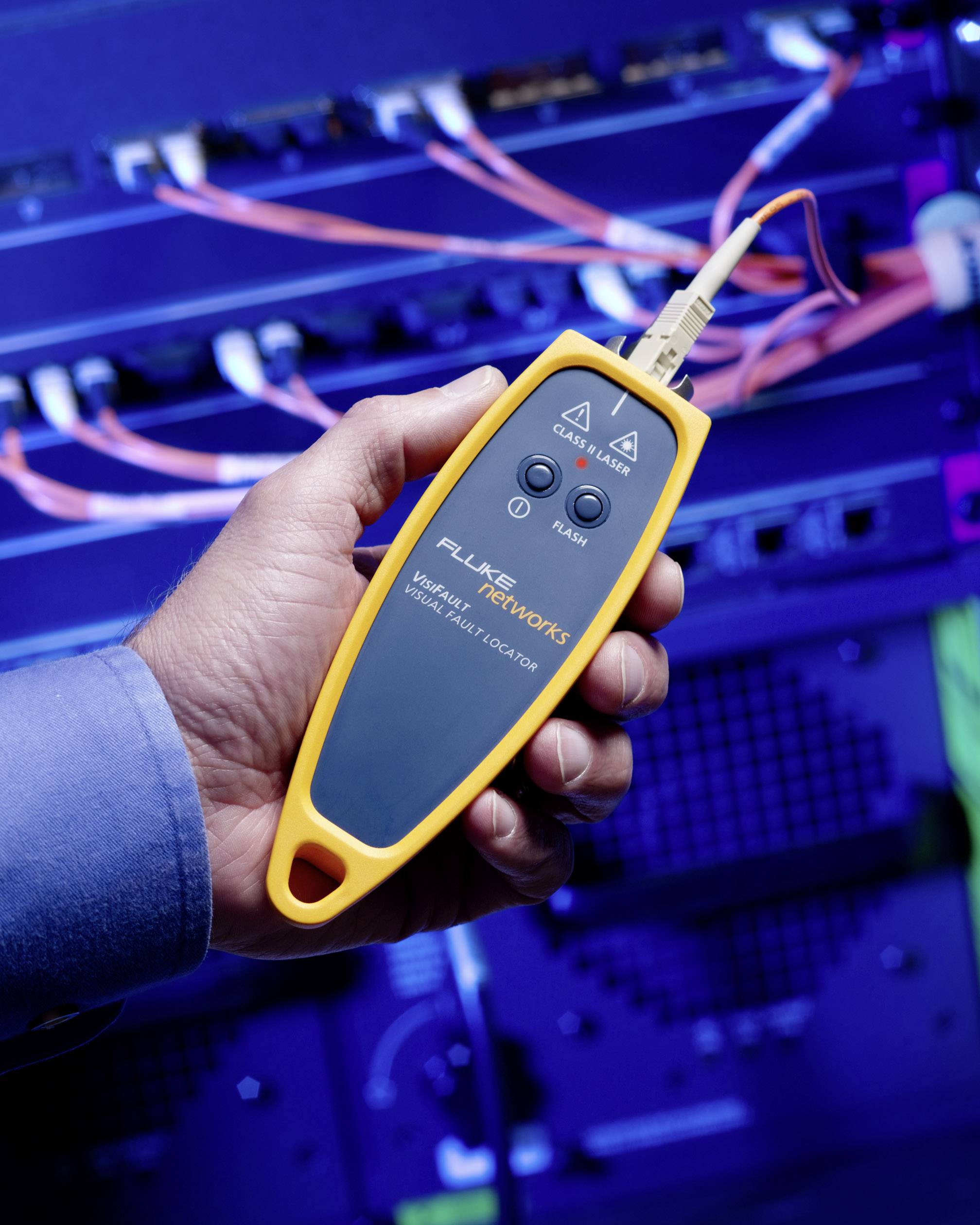 'A hand holds a yellow cable tester in front of a server with orange and blue cables.'