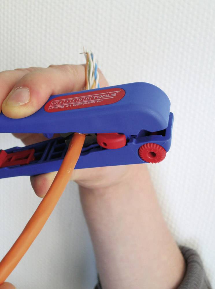A hand is holding a cable tool stripping an orange cable. A white wall is visible in the background.