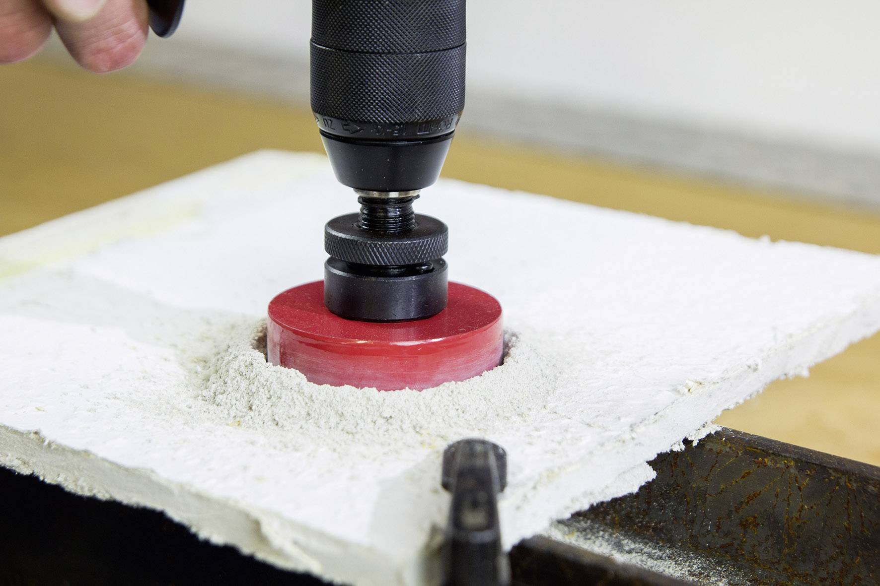 A hand is holding an electric drill with a rotating drill bit, drilling a round hole into a white, plasterboard surface.