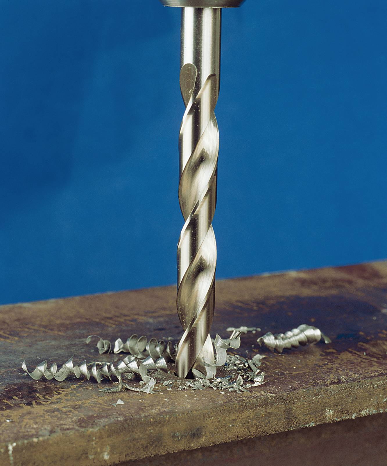 A drill penetrates a metal surface, surrounded by metal shavings. The background is blue, with emphasis on the drilling action.