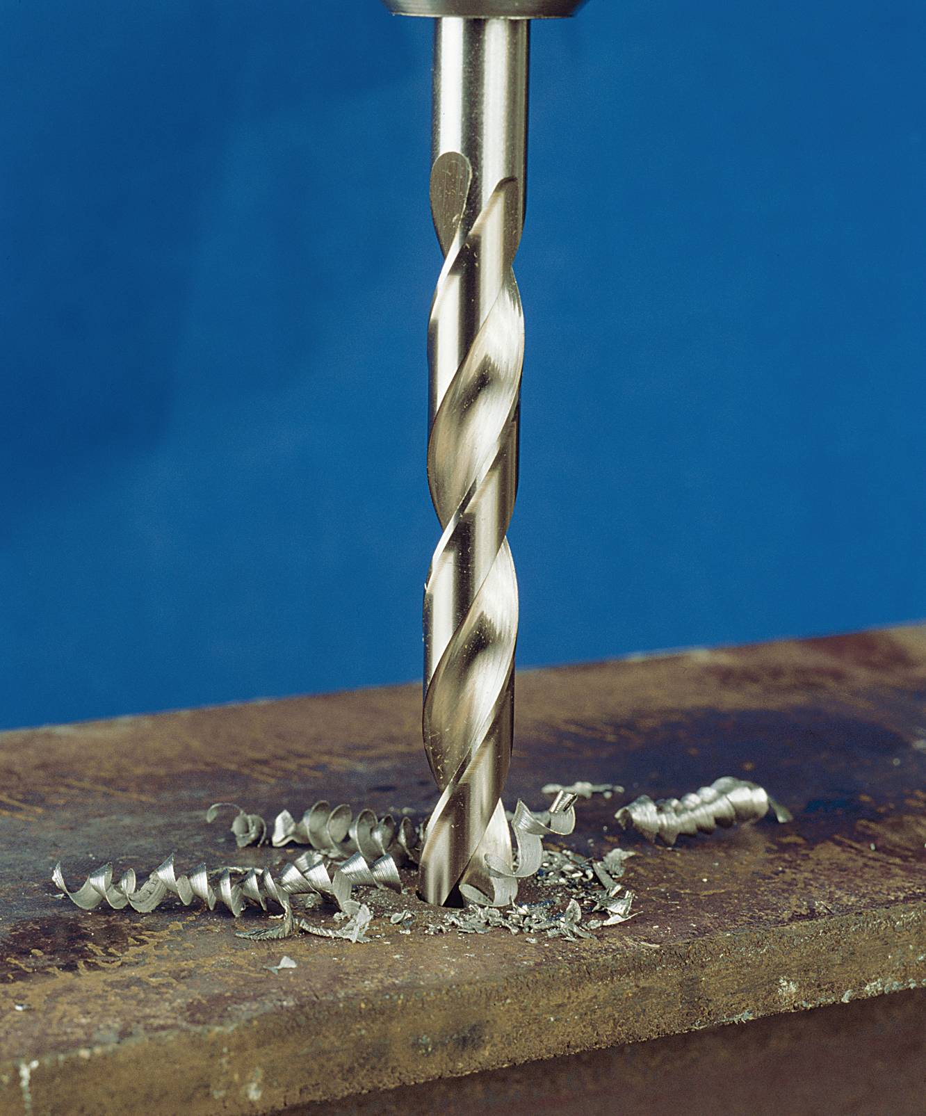 A metal drill pierces a metal plate, creating spirals of metal shavings. The background is blue.
