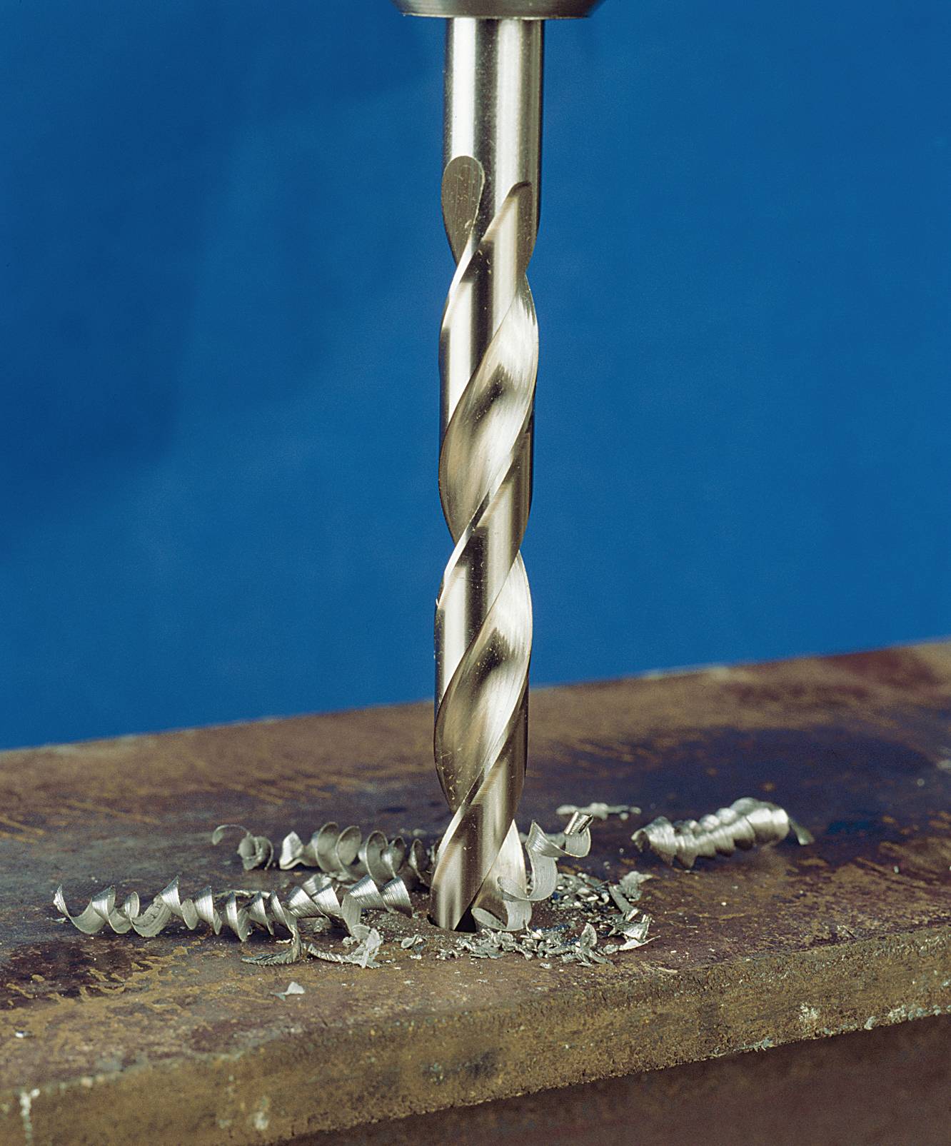 A metal drill is drilling a hole in a metal plate, surrounded by metal shavings, against a blue background.