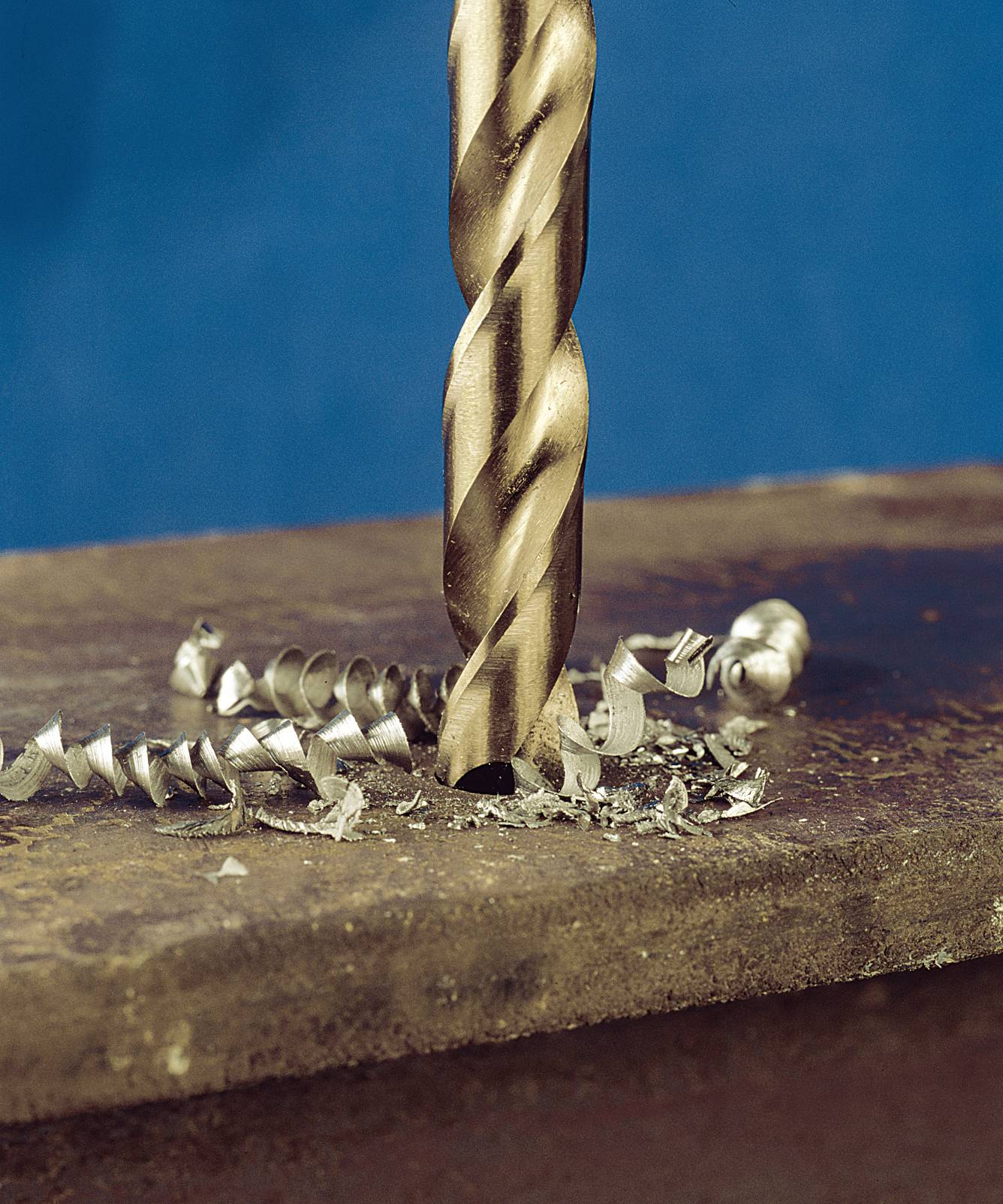 A metal drill bit pierces a metal plate, surrounded by metal shavings.