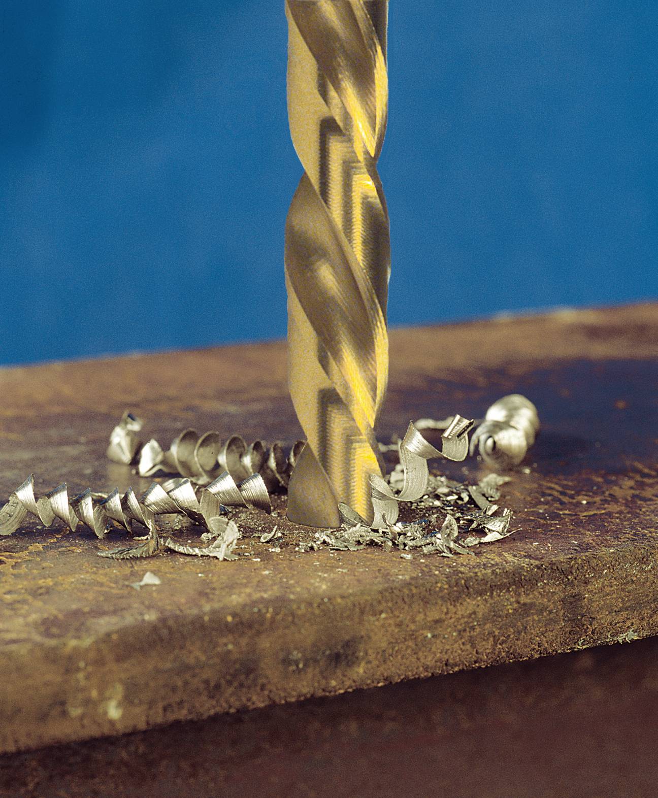 A metal drill is drilling into a metal plate. Metal shavings are scattered around the drill. The background is blue.