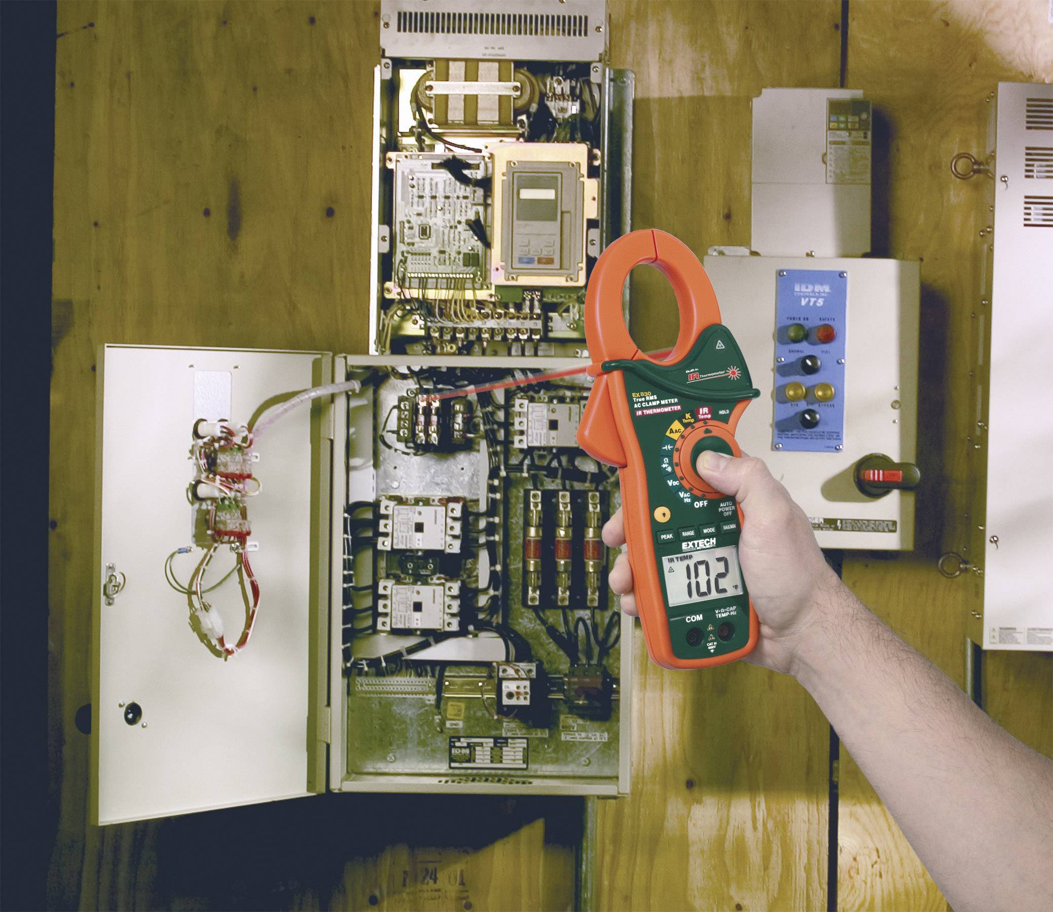 A person is holding an ammeter showing 10.2, standing in front of an open electrical cabinet with multiple cables and electronic components.