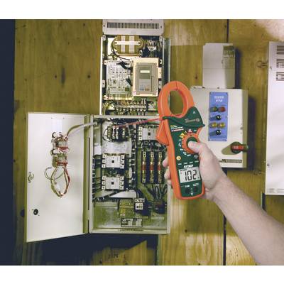 A person is holding an ammeter showing 10.2, standing in front of an open electrical cabinet with multiple cables and electronic components.