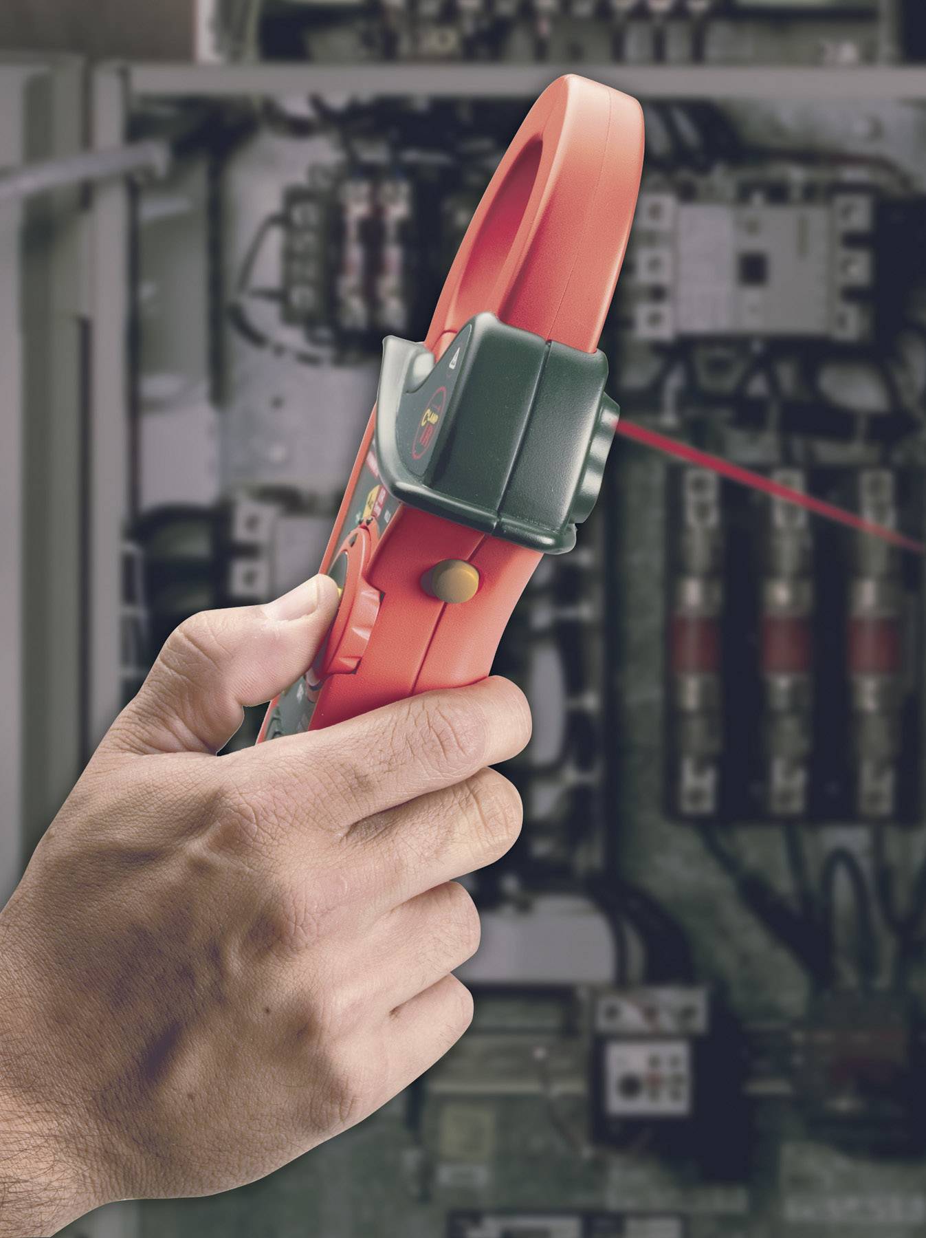 A hand holds a red electrical measuring device in front of a switchboard with cables and fuses. The device is measuring voltage.