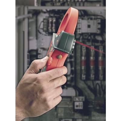 A hand holds a red electrical measuring device in front of a switchboard with cables and fuses. The device is measuring voltage.