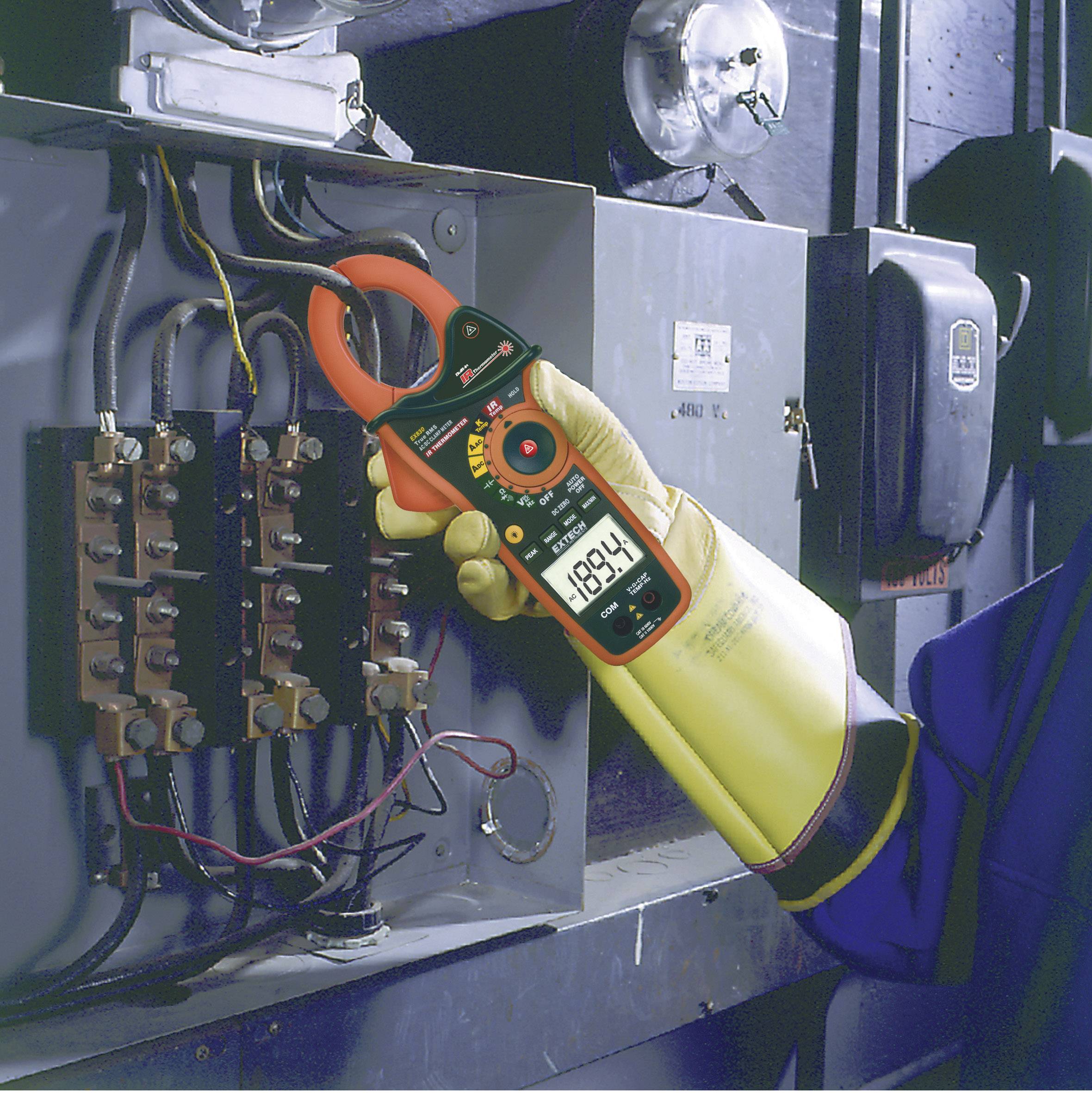 A person is wearing yellow protective gloves and holding a clamp multimeter to carry out electrical measurements on an electrical cabinet.