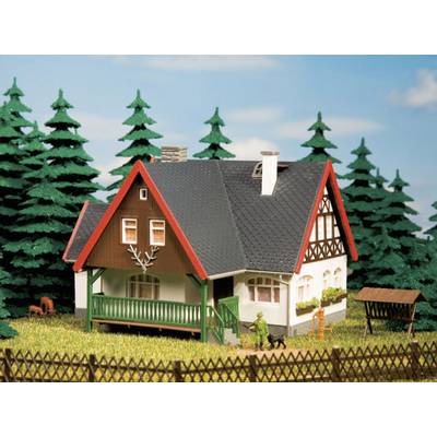 A small model house with a gabled roof, veranda and half-timbered details stands surrounded by pine trees and a wooden fence in a rural setting.