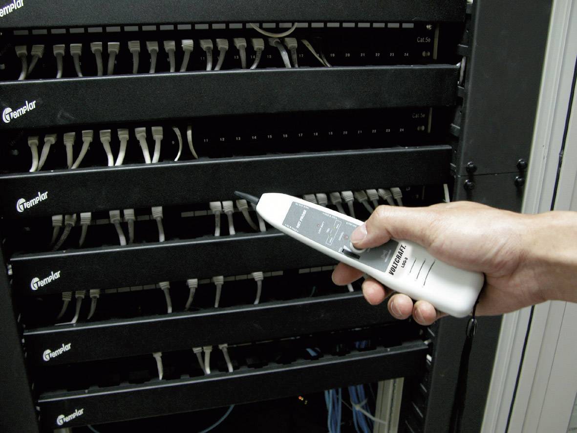 A hand holds a cable testing device in front of a server cabinet full of cable connections.