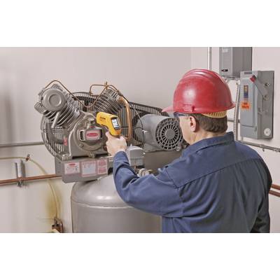 A man in a hard hat and work attire is checking an industrial compressor with an infrared thermometer.