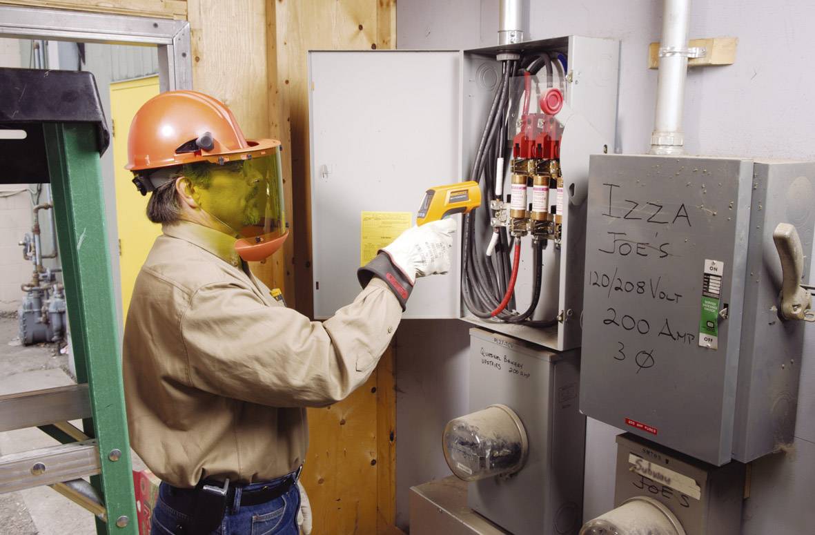 A person wearing a protective helmet and face shield is measuring the temperature of an electrical switchboard using an infrared thermometer.
