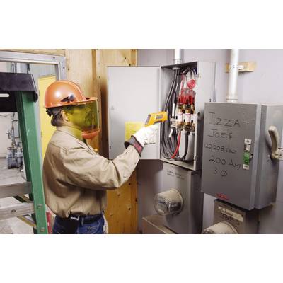 A person wearing a protective helmet and face shield is measuring the temperature of an electrical switchboard using an infrared thermometer.