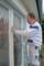 A man in workwear is standing on a ladder and applying protective plastic sheeting to a window frame, presumably in preparation for painting.