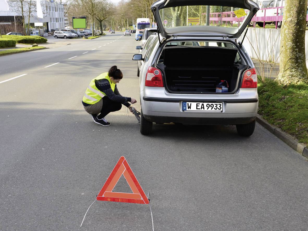 A woman in a high-visibility vest is changing a car tyre on a road with the boot open. A red warning triangle is set up.
