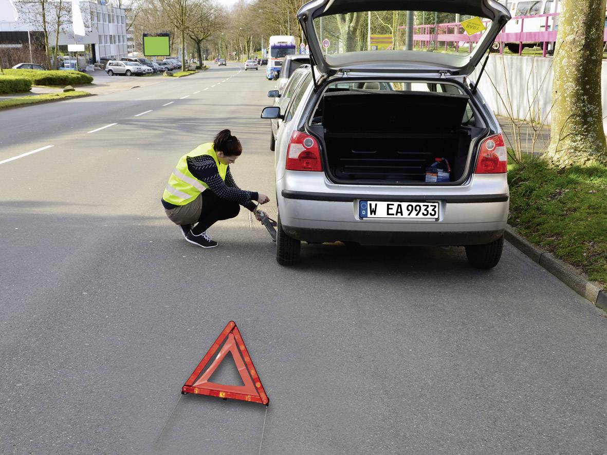 A woman is changing a tyre on a car by the roadside with the boot open. A warning triangle is set up.