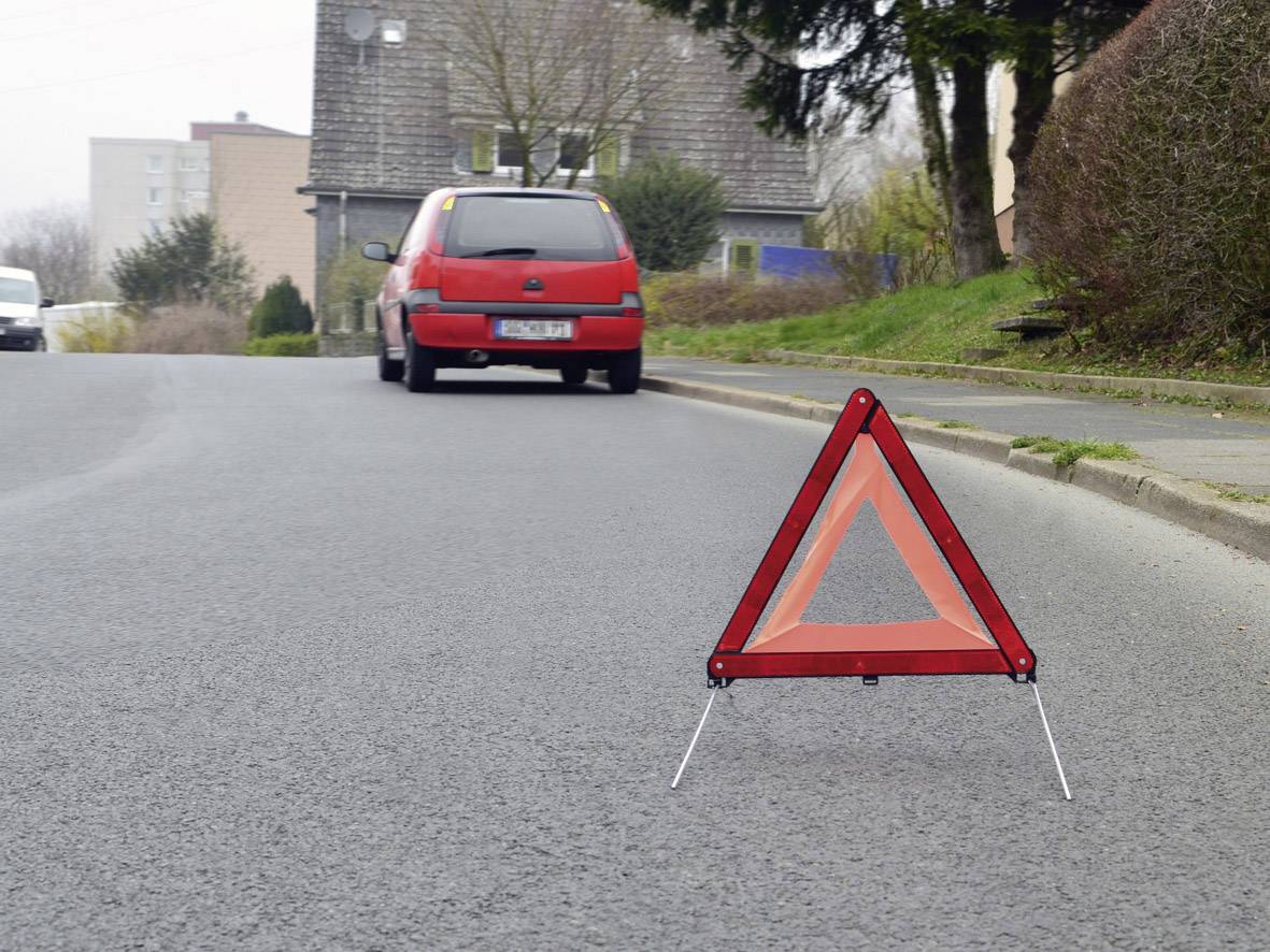 A warning triangle is standing on a road, with a red car parked in the background. The scene suggests a possible breakdown.
