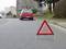 A warning triangle is standing on a road, with a red car parked in the background. The scene suggests a possible breakdown.