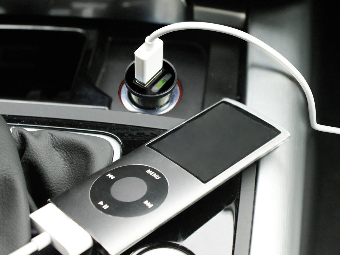 A portable music player is being charged in the car via an adapter. The device has a rectangular display and touch buttons.