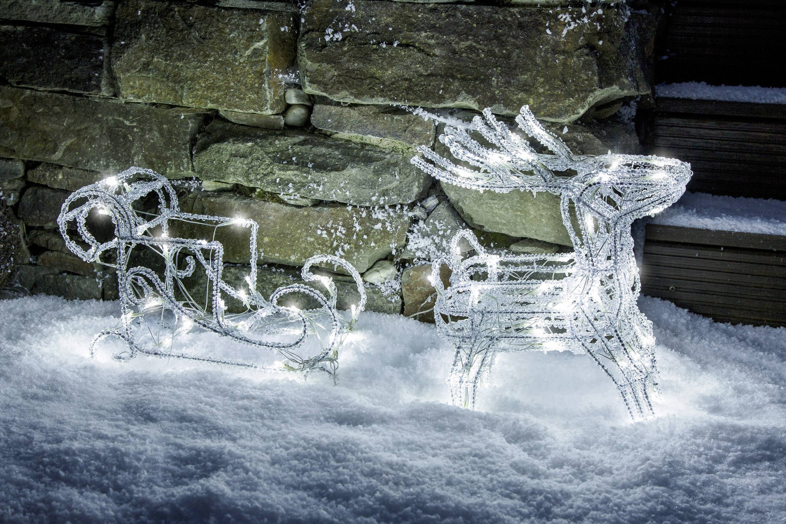 A luminous reindeer and sleigh frame made of wire on snow-covered ground in front of a stone wall.