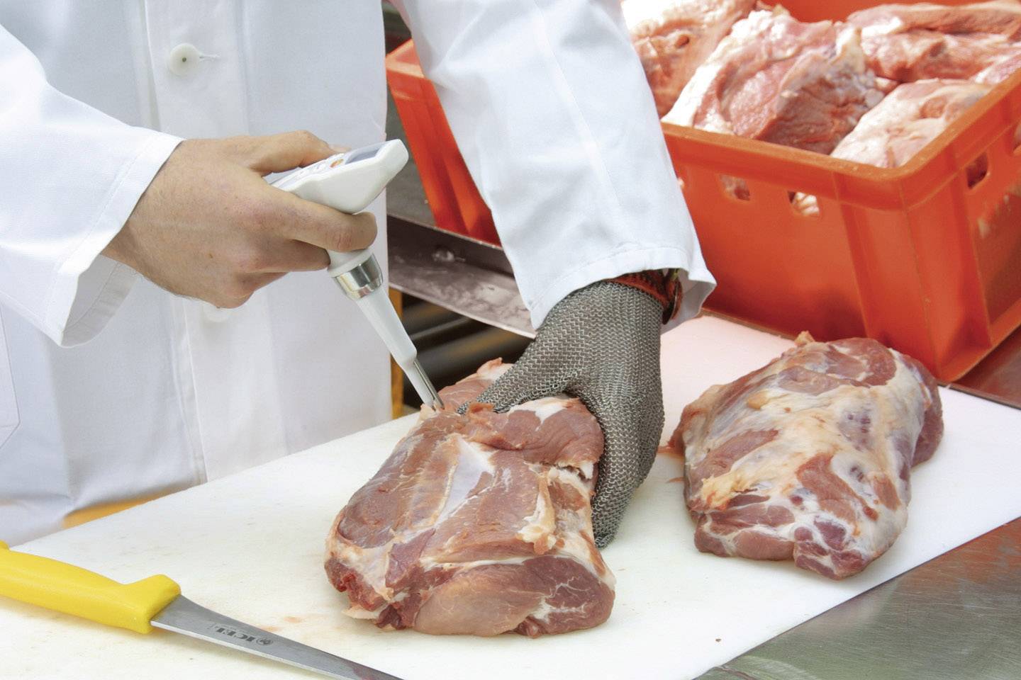 'Person in a white coat sprays liquid onto raw meat on a chopping board. Pieces of meat in a red crate are visible in the background.'