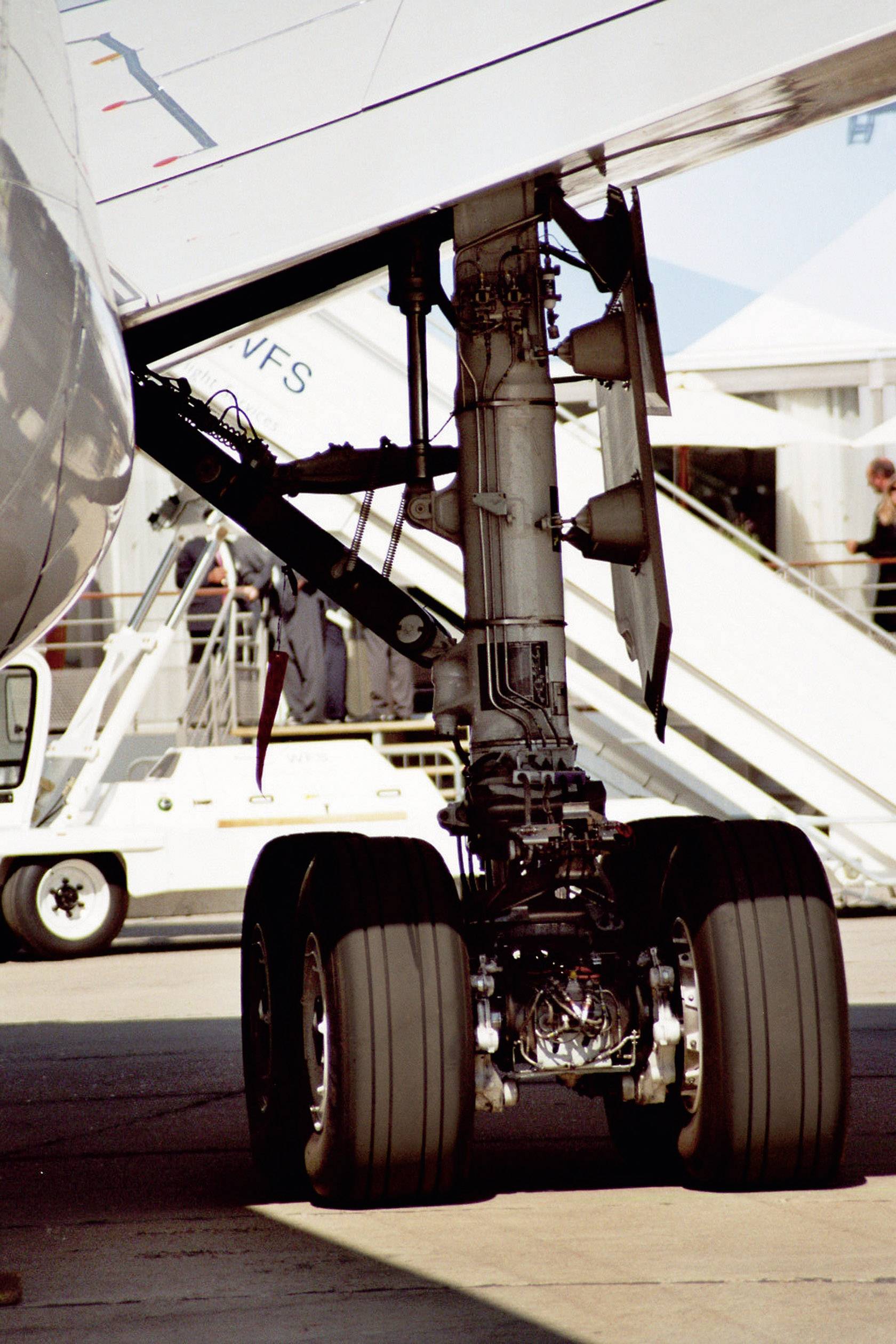 The image shows the landing gear of an aircraft on the ground. In the background, stairs and a building are visible.