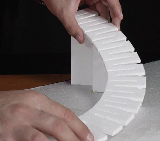 Two hands are holding a curved polystyrene strip shaped like a staircase, balancing it on a white block.